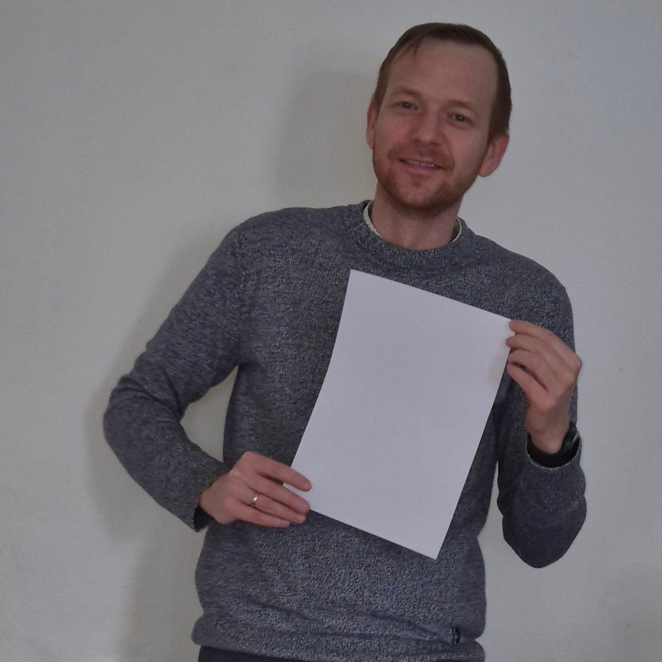 Man in een grijs trui houdt een leeg vel papier vast en glimlacht.