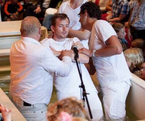 Een man wordt gedoopt in een baptisterium door twee anderen, met publiek op de achtergrond.